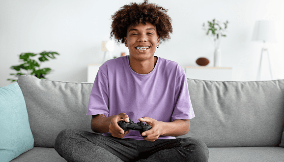 Young person with curly hair in a purple shirt sitting cross-legged on a gray sofa, smiling while holding a video game controller, in a bright room.