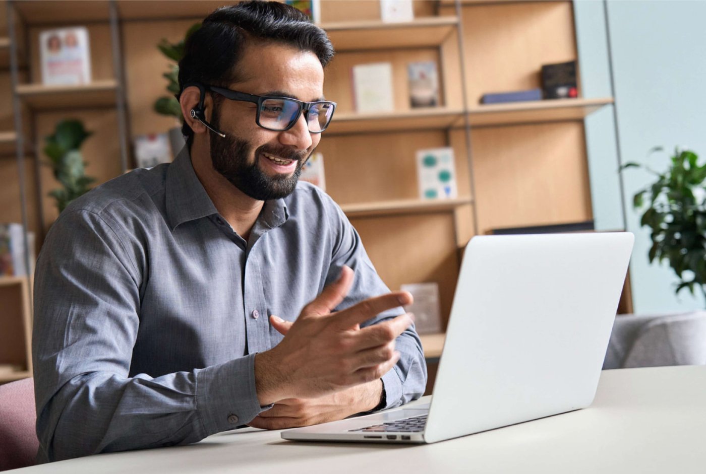 A smiling man with glasses and a headset gestures during a video call on a laptop. He is seated in a modern office with shelves and plants behind him.