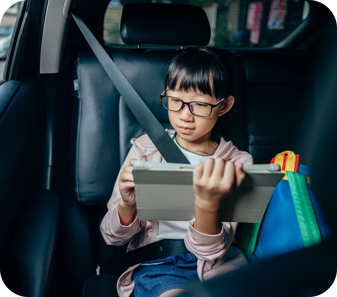Young girl wearing glasses focused on a tablet in a car's backseat. She's buckled in and a colorful backpack is next to her, conveying concentration.